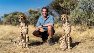 Een foto van bioloog Freek in Afrika. Hij zit gehurkt op de grond en glimlacht naar de camera. Links en rechts van hem zit een cheeta.