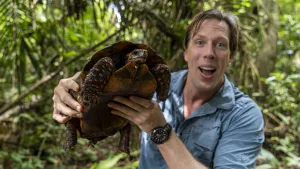 Een foto van bioloog Freek. Hij houdt een schildpad vast en praat in de camera. Op de achtergrond zie je allemaal tropische bomen en planten.