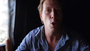 Een foto van bioloog Freek. Hij praat naar de camera. Freek zit in een schuilhut zo te zien. De achtergrond is heel donker, maar links is een opening waardoor er licht naar binnen komt.