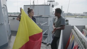 Een foto van een presentator en Lisa op een Nederlands schip. Lisa heeft een grote vlag vast die schuin opgedeeld is in twee kleuren: geel en rood. De presentator leunt op een hek waar tientallen vlaggen achter hangen voor het versturen van een boodschap.