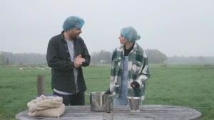 Een foto van Janouk en een man op een grasveld die een blauw netje op hun hoofd hebben. Voor hen staat een tafel met daarop een blauw bakje achter een witte zak met niet-zichtbare sojabonen, een pan met plastic staafmixer erin en een klein emmertje.