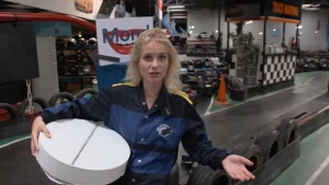 Een foto van Janouk in het midden van een kartbaan binnen met op de achtergrond karts en helmen. In haar rechterarm heeft ze een hele grote, ronde en witte pil met in het midden een gleuf, precies zoals een aspirine pil. Ze draagt een zwart met blauw pak.