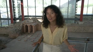 Een foto van Serah die voor een stalen hek staat. Achter haar zijn afgebrokkelde stukken van een muur zichtbaar en opgestapelde stapeltjes kleine, bruine stenen met drie grotere stukken steen erop. Dit is een opgegraven gedeelte van een Romeins badhuis.