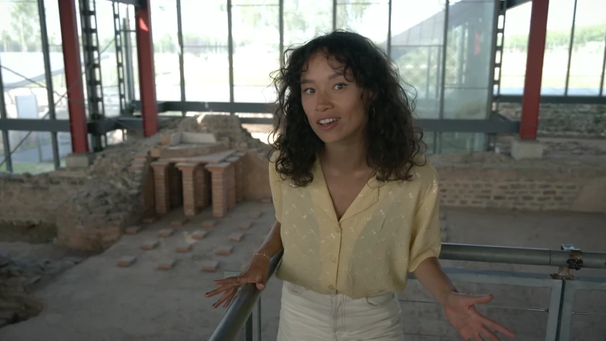 Een foto van Serah die voor een stalen hek staat. Achter haar zijn afgebrokkelde stukken van een muur zichtbaar en opgestapelde stapeltjes kleine, bruine stenen met drie grotere stukken steen erop. Dit is een opgegraven gedeelte van een Romeins badhuis.