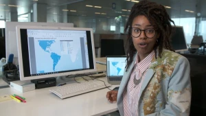 Een foto van presentator Eva in een jasje met de wereldkaart erop. Op het bureau achter haar staat een beeldscherm en een laptop waarop een blauwe kaart staat met daarop alle ambassadeurs van Nederland in de Verenigde Staten van Amerika en Zuid-Amerika. 