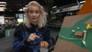 Een foto van Janouk in een zwart en blauw pak naast een lichtbruin bord met gaatjes. Het bord kan tonen hoe een seintje van pijn doorgegeven wordt. Op een groen getekende hand kan iets vallen in een buisje, vallen op een bakje en een aan-uit knop raken.