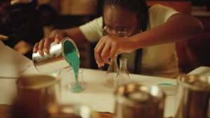 Een foto van presentator Tirsa die een blik met lichtgroene verf in een kleine glazen maatbeker voor haar op een tafel giet. Er staan ook twee andere, lege glazen flesjes op met een smalle bovenkant en bredere onderkant.  