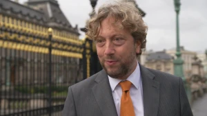 Een foto van de Nederlandse ambassadeur Pieter Jan Kleiweg die naast het hek van het Koninklijk Paleis in België staat. Hij draagt een grijs pak met een witte blouse en een lichtbruine stropdas. Het hek  naast Pieter Jan is zwart met goudkleurige punten.