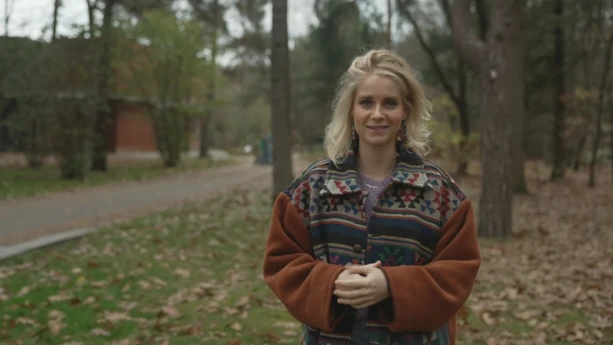 Een foto van presentator Janouk. Ze staat in een bos en kijkt in de camera. Op de achtergrond zie je een gebouw. 