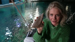 Een foto van Janouk die naast een groot aquarium, een basin, in de dierentuin in Arnhem zit. In haar rechterhand houdt ze een bruingrijze rugvin van een haai omhoog naar de camera. Janouk kijkt in de camera. Ze draagt een groene trui en lichtblauwe broek.