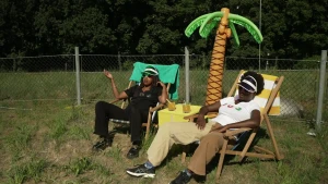 Een foto van presentatoren Eva en Tirsa die op een klapstoel zitten op een grasveld naast frisdrank op een tafel en een opblaaspalmboom. Ze dragen een zonnebril en een zonneklep. Eva draagt zwarte kleding, Tirsa draagt een wit shirt en beige broek.