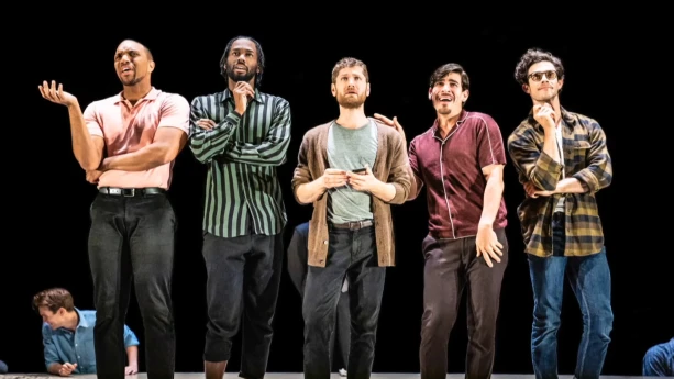 Een foto van vijf mannen die naast elkaar op een podium staan. Een paar van hen lijken iets te zeggen. De achtergrond is zwart van kleur.