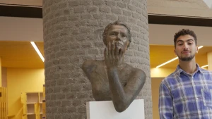 Een foto van presentator Nizar die naast een standbeeld van Marga Klompé staat. Marga houdt haar hand met de vingers omhoog een stukje van haar gezicht af met de handpalm naar het gezicht toegericht. Ze was in 1956 als eerste Nederlandse vrouw minister.