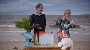 Een foto van Janouk en een man op het strand achter een tafel met een bord waar bovenaan rood en eronder blauw vocht zit, links een blauw bord "Bonaire", een palmboom en een fles met bericht, rechts een rood bord "Noordpool" en een ijsbeerknuffel. 