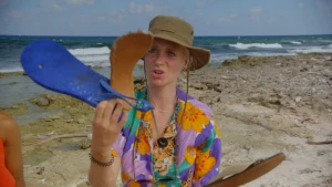 Een foto van Janouk op het strand van Bonaire die twee helften van een kapotte, gevonden slipper voor de camera houdt. Ze is plastic aan het opruimen. Janouk heeft een blouse met oranje en paarse bloemen aan en een bruine hoed met een koort op.