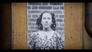Een zwart-wit foto van Truus van Lier, een Joods-Nederlandse vrouw die tijdens de Tweede Wereldoorlog in het verzet zat. Ze staat voor een muur van bakstenen en heeft een donkere jurk aan met witte bloemetjes erop. De foto hangt op een houten achtergrond.