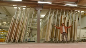 Een foto van presentator Pascal een grote hal in een fabriek. Hij staat voor een groot metalen rek met daarin houten muren. Er staat een grote metalen kolom in het midden. 