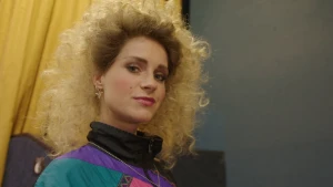 Een foto van presentator Janouk met jaren tachtig kleding en make-up. Ze draagt een trainingsjasje, heeft een grote bos blonde krullen en veel oogschaduw op.