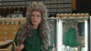 Een foto van Janouk die een pruik opheeft met bruin haar en lange krullen. De krullen staan omhoog op haar hoofd en er lopen krullen langs beide kanten van haar gezicht naar beneden. Ze zit voor een verlichte spiegel met twee schuine kanten.