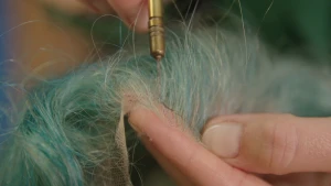 Een foto van een hand die een pruik met blauw haar vasthoudt. De andere hand houdt een stuk gereedschap tegen de pruik aan. Het is een metalen handvat met daaraan een dunne kromme naald.