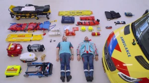 Een foto van 2 mensen die recht naast elkaar op de grond liggen. Ze hebben speciale kleding aan voor op de ambulance. Om hen heen liggen allerlei voorwerpen netjes naast elkaar gelegd. Heel veel dingen zijn fel geel of rood. Naast hen staat een ambulance.
