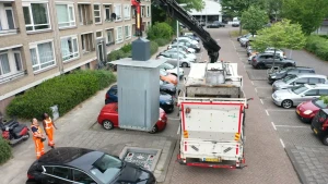 Een foto van een straat met een vuilniswagen. Een hijskraan op de wagen tilt een ondergrondse afvalbak uit de stoep. Dat is een grote grijze bak. Links op de foto staat de presentator Tirsa met een vuilnisman. Rondom staan huizen en auto's.