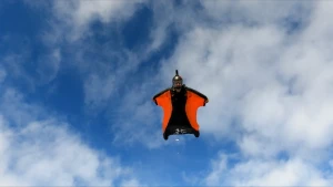 Een foto van iemand in een wingsuit in de lucht. Een wingsuit is een speciaal pak met vleugels, waardoor je blijft zweven. De vleugels zijn een stuk stof tussen de benen en tussen de armen en de benen.