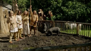 Een foto van een groep mensen verkleed alsof ze uit de oertijd komen. Ze hebben bijlen en ander gereedschap in de hand. Ze staan te juichen bij een stukje kale grond met een varken. Achter hen staan tenten, een houten hek en een bos.