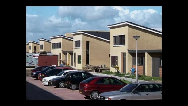 Een foto van een straat met nieuwe huizen. Ze zien er allemaal hetzelfde uit. Voor die huizen zijn parkeerplaatsen met auto's. 