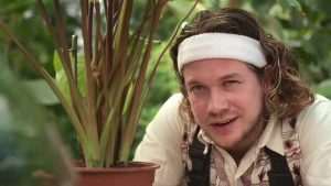 Een foto van presentator Pascal die naast een plant staat. Van de plant die in een rode plastic pot staat, zijn alleen de groene stengels te zien. Pascal heeft een witte zweetband om zijn hoofd en vertelt over de zwetende bladeren van de plant.