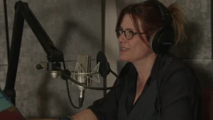 Een foto van een vrouw achter een microfoon. Ze heeft een grote koptelefoon op. Ze zit vlak bij een muur met grote grijze platen erop.