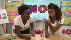 Een foto van presentatoren Tirsa en Eva die achter een tafel staan. Op de tafel staat een glazen vaas waarin op een laag water een rode appel ligt. De appel drijft in het water, voor de helft boven en voor de helft onder het streepje dat op de vaas staat.