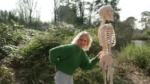 Een foto van de presentator en een skelet die buiten staan tussen wat struiken. De presentator kijkt naar de camera en houdt een hand bij de onderkant van haar rug. Haar andere hand zit op de heup van het skelet.
