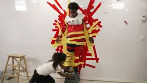 Een foto van Tirsa die tegen een witte muur geplakt staat met rode en gele tape. Tirsa staat op een klein grijs krukje en kijkt naar het krukje. Eva zit op haar knieën op de grond en heeft het krukje vast met beide handen. 