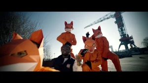 Een foto van vier mensen die buiten staan met een oranje werkpak aan, zwarte vingerloze handschoenen aan en een masker van een vos op hun hoofd. Middenin staat rapper Leafs. Op de achtergrond staat een hijskraan.