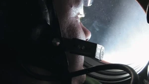 Een foto van het gezicht van astronaut André Kuipers dat van achteren zichtbaar is. Hij staat bij een klein, hol en rond raam naar buiten te kijken de ruimte in. Hij heeft een headset op zijn hoofd waarvan de microfoon naast zijn mond zichtbaar is.