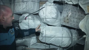 Een foto van astronaut André Kuipers met een heleboel witte afvalzakken. De zakken zitten achter een dun metalen hek. André kijkt en wijst naar de zakken.