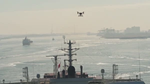 Een foto van een drone die boven een schip vliegt. Een drone is een heel kleine helicopter of vliegtuig, waarmee je op afstand iets kunt doen. Op de achtergrond is veel water, meer schepen en een haven.