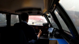 Een foto van een treinmachinist in de bestuurdersruimte van de trein. De machinist kijkt voor zich door de ruit en links en rechts in de ruimte zijn ook ruiten te zien. Hij zit voor knopjes, metertjes, een lichtje, een hendel, een tablet en een camera.