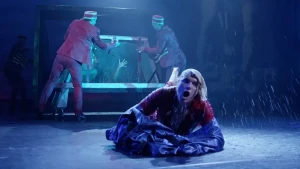 Een foto van Goochelaar en illusionist Hans Klok die kletsnat en happend naar adem op een blauwe doek op de vloer ligt. Hij is uit een bak met water gevallen die op de achtergrond is opengedaan door twee mannen in een rood kostuum met rood rond petje.  