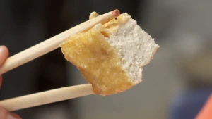 Een foto van een blokje tofu dat met twee houten eetstokjes voor de camera gehouden wordt. Het blokje heeft een goudgele korst en een witte binnenkant. Tofu wordt gemaakt van blokjes soja en kan daarom gegeten worden bij een vegetarische maaltijd. 