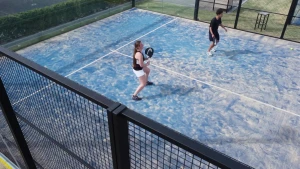 Een foto van Nizar en Tess van Dinteren, die op een blauwe baan met witte lijnen padel spelen. Bij padel sla je de bal met een racket tegen een glazen wand en dan slaat je tegenstander de bal terug. Nizar wacht tot de bal terugkomt van de glazen wand.