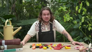 Een foto van Pascal met achter zich groene planten en voor zich op tafel pepers die rood, geel of groen van kleur zijn. Links op de tafel staat een gele gieter op een stapel van drie boeken en rechts op tafel liggen een klein tuinharkje en tuinschepje. 