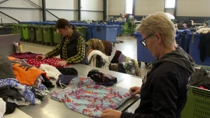 Een foto van twee medewerkers van het Leger Des Heils die op een tafel tweedehands kleding controleren. Op de foto bekijkt iedere medewerker een blouse om te bepalen hoe geschikt de blouse is om opnieuw te dragen. Achter hen staan grote verzamelbakken.