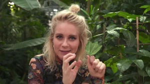 Een foto van presentator Janouk met een groen blad in haar linkerhand. Ze houdt de vingers van haar rechterhand gekromd omhoog onder haar gezicht en kijkt recht in de camera. Achter Janouk staan allemaal groene planten met kleine en grotere bladeren.