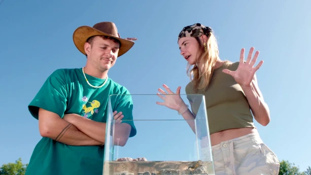 Een foto van Jurre en Emma die buiten staan. Voor hen staat een glazen bak met een Geelbuikvuurpad erin. Emma is iets aan het vertellen aan Jurre. Jurre kijkt naar de glazen bak.