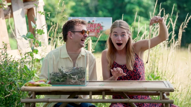 Een foto van Jurre en Emma die buiten aan een tafel zitten. Op de tafel staat een glazen bak met planten erin. Emma houdt een slangenhuid in de lucht en kijkt verbaasd. Jurre lacht naar haar.