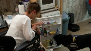 Een foto van een vrouw die in haar rolstoel in de keuken zit. Ze heeft de koelkast openstaan. Naast de koelkast staat haar zwarte hulphond, een hond die speciaal getraind is om haar te helpen. De hond, die Noemie heet, pakt een flesje uit de koelkast.