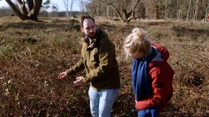 Een foto van presentator Janouk en ecoloog Henrik Barmentlo. Een ecoloog weet veel over natuurlijke leefgebieden en soorten dieren en planten. Ze staan in een open veld en Henrik wijst met zijn handen naar een struik met een paar bladeren aan de takken