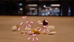 Een onscherpe foto van etensresten die op de grond liggen, zoals een stukje bloemkool, een spekje en een bruin nootje. Voor de foto zijn allemaal bacteriën getekend. Ze hebben een rode rand en roze binnenkant, met rode rondjes en zwarte streepjes erin.